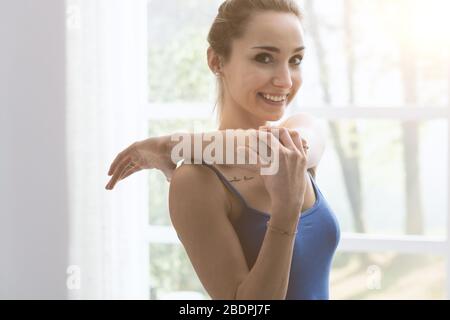 Giovane donna sportiva che si esercita a casa nel soggiorno, si stende il braccio e sorridente alla macchina fotografica Foto Stock