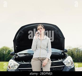 Giovane donna triste che chiama un servizio di riparazione auto con il suo telefono, sta avendo un guasto auto in una strada di campagna Foto Stock