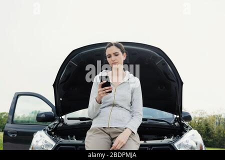 Giovane donna triste che chiama un servizio di riparazione auto con il suo telefono, sta avendo un guasto auto in una strada di campagna Foto Stock