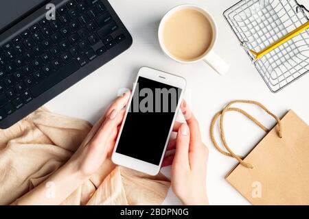 Concetto di shopping online su cellulare. Vista dall'alto mani femminili che tengono lo smartphone sopra il tavolo con computer portatile, tazza da caffè, carrello, coperta Foto Stock