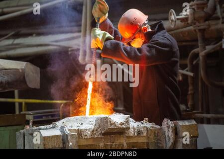 lavoratore con elmetto e maschera viso agita metallo liquido in un forno da barra di acciaio Foto Stock