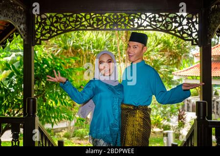 Un ritratto di giovane coppia di musulmani malesi in costume tradizionale durante la celebrazione di Aidilfitri mostrando gesto di benvenuto da casa tradizionale di legno. R Foto Stock