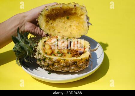 Riso fritto con ananas con gamberi in ananas su piatto bianco con sfondo giallo, contenitore per alimenti eco-friendly Foto Stock