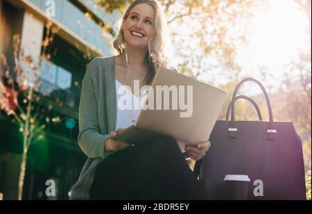Donna che indossa auricolari seduti all'esterno e che utilizza un computer portatile. donna d'affari seduta all'aperto in città con un laptop in una giornata di sole. Foto Stock