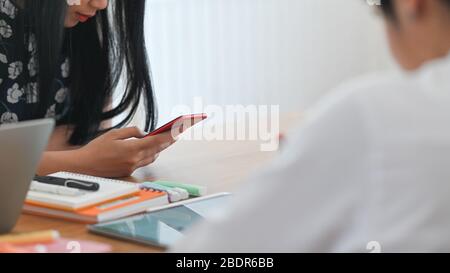 Immagine ritagliata delle mani di una giovane donna creativa che tiene uno smartphone con schermo vuoto mentre si è seduti alla scrivania sopra un comodo soggiorno come Foto Stock