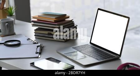 Computer portatile a schermo vuoto che mette sul tavolo di lavoro con una pila di libri, una tazza da caffè, appunti, un tablet per computer, una lente di ingrandimento. Ambiente di lavoro ordinato Foto Stock