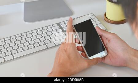 Un'immagine tagliata di smart man che tiene uno smartphone bianco con schermo nero vuoto in mano mentre si è seduti al moderno tavolo da lavoro con comoda scrivania Foto Stock