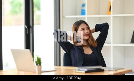 Donna rilassante dopo aver finito il lavoro mentre si siede alla moderna scrivania di fronte al suo computer portatile e libri con libreria bianca moderna e vincere Foto Stock