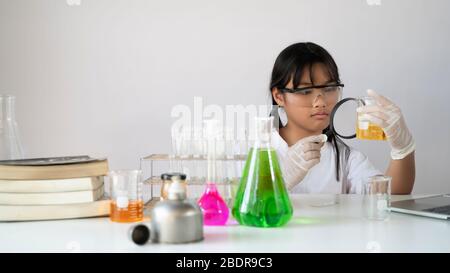 Foto di adorabile scolaretta che guarda attraverso una lente di ingrandimento mentre fa un esperimento scientifico con vetreria chimica sopra il laboratorio bianco Foto Stock