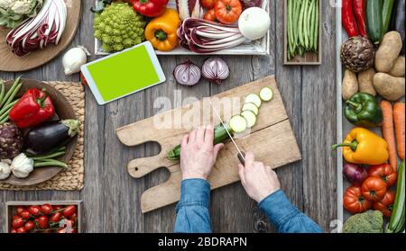 Uomo che cucina e affetta verdure fresche su un piano di lavoro rustico cucina, sta cercando le ricette online utilizzando un tablet digitale Foto Stock