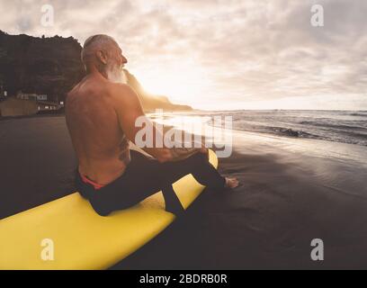 Uomo anziano felice di idoneità che siede sul surfboard che guarda il tempo del tramonto - surfista maturo bearded che si diverte il giorno di surf Foto Stock