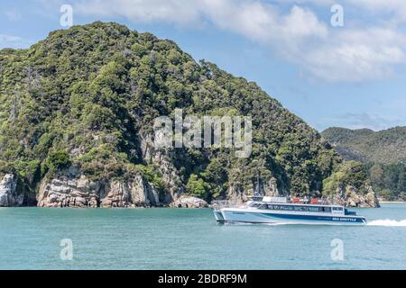 KAITERITERI, NUOVA ZELANDA - Novembre 15 2019: Paesaggio con nave da passeggeri romantici vela vicino verde promontorio roccioso a Awaroa Bay, girato in Bright spr Foto Stock