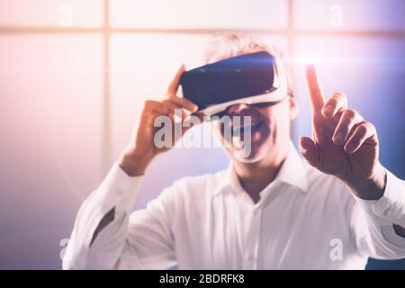 Uomo maturo e sicuro che indossa uno spettatore VR e interagisce con un'interfaccia utente per la realtà virtuale, punta con un dito Foto Stock