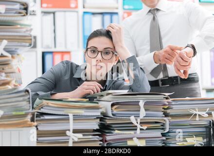 Il boss esigenti rivolta verso il suo orologio e chiedendo al suo dipendente per affrettare il suo segretario è frustrato e sopraffatta dal lavoro: business e deadlin Foto Stock