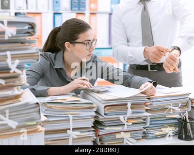 Il boss esigenti rivolta verso il suo orologio e chiedendo al suo dipendente per affrettare il suo segretario è frustrato e sopraffatta dal lavoro: business e deadlin Foto Stock