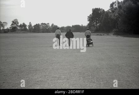 anni '60, storico, su un campo da golf, in una linea, tre uomini che camminano lungo un fairway, due golfisti e un caddie che sta portando i randelli per uno di loro e che sta portando un vestito, mentre l'altro golfista sta tirando una borsa da golf su un carrello dell'epoca. Foto Stock
