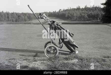 Anni '60, storico, all'esterno su un campo da golf, seduto su una tee, una borsa da golf in pelle con mazze su un carrello da golf a ruote metalliche dell'epoca. Foto Stock