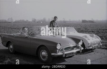 Anni '60, storico, automobilistico, due auto sportive open-top 2 porte dell'epoca parcheggiate l'una accanto all'altra accanto a un campo di colture, vicino a Eastholm Green, Letchworth Garden City, Herts, Inghilterra, Regno Unito. Una coppia matura è seduta nella loro Sunbeam Alpine, con il Signore che fuma un tubo. Un uomo più giovane, forse il figlio, è seduto in un Austin Healey Sprite MK1, noto come 'Frogeye' a causa dell'aspetto dei fari delle auto sul cofano. Foto Stock