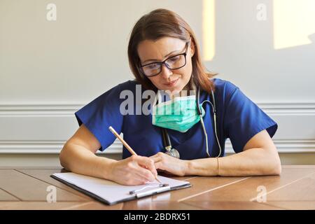 Medico femminile in uniforme blu, maschera medica seduta a tavola, scrittura sulla clipboard Foto Stock