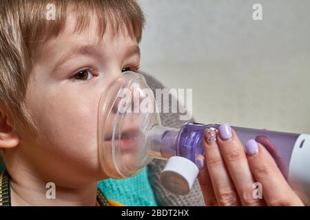 Un ragazzino che soffre di malattia asma bronchiale che riceve il trattamento respiratorio dalla madre con inalatore aerosol. Trattamento delle complicanze di vi Foto Stock