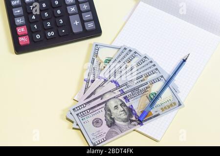 Diverse nuove banconote da cento dollari si trovano su un pad bianco vuoto. Smartphone, calcolatrice e penna su tavolo giallo Foto Stock