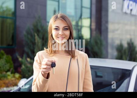 Ritratto closeup felice, sorridente, giovane donna attraente, compratore vicino alla sua nuova auto mostrando le chiavi isolate fuori del concessionario, l'ufficio del lotto della concessionaria. Personale Foto Stock