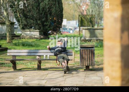 Un uomo seduto su una panchina in un piccolo parco e bere sidro, gode il sole primaverile, Inghilterra, Regno Unito Foto Stock