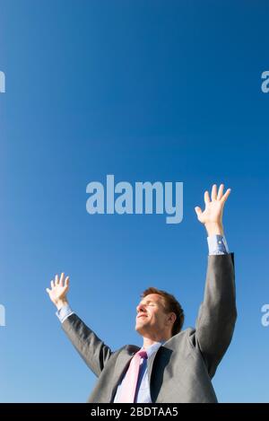 Uomo d'affari in piedi all'aperto tenendo le braccia in alto di fronte al sole contro il cielo blu chiaro Foto Stock
