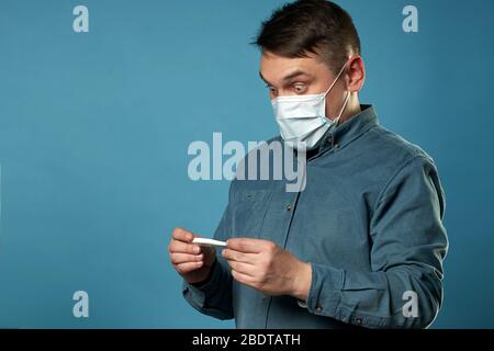 Giovane uomo in tessuto casual indossare maschera medica protettiva eccitato, stressato e spaventato dopo la lettura molto alta temperatura risultato da digitale Foto Stock