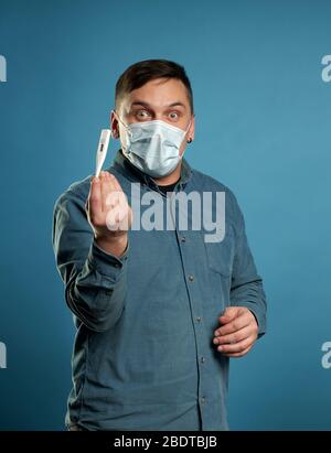 Giovane uomo in tessuto casual indossare maschera medica protettiva eccitato, stressato e spaventato dopo la lettura molto alta temperatura risultato da digitale Foto Stock