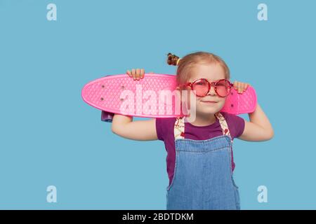 Piccola bambina fresca in abiti luminosi siede su uno skateboard e guardando la macchina fotografica isolato su sfondo blu Foto Stock