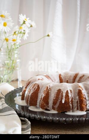 Torta al limone con fiocco di ricotta e glassa, esposta su un piatto decorativo nero con fiori Daisy in un vaso sullo sfondo. Foto Stock