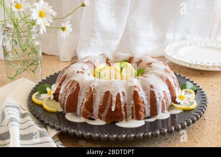 Torta al limone con fiocco di ricotta e glassa, esposta su un piatto decorativo nero con fiori Daisy in un vaso sullo sfondo. Foto Stock