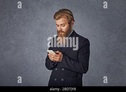 Uomo d'affari bearded che tiene guardando il telefono che testing isolato grigio parete di fondo Foto Stock
