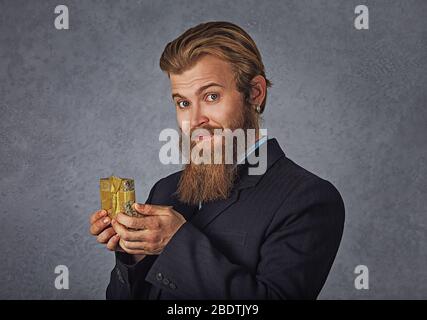 Bearded eccitato uomo che si apre guardando dono d'oro isolato su parete di fondo grigia. Concetto di vacanza invernale Foto Stock