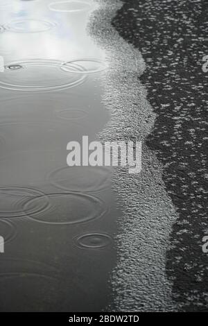 Dove un bordo di una pozzanghera incontra un sentiero nero asfaltato, le gocce cadono nell'acqua e sull'asfalto, creando un astratto surreale di molte texture Foto Stock