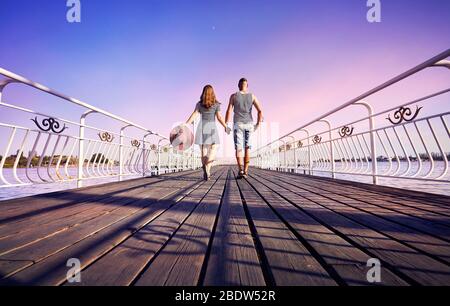 Donna in abito a righe con Red Hat e Uomo in camicia strippato andando sul molo vicino al lago al tramonto rosa Foto Stock