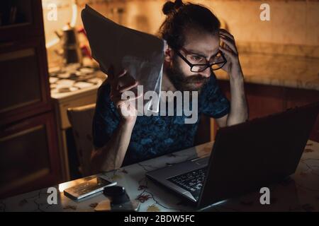 Stressato giovane hippster lunghi capelli bearded uomo sta controllando la fattura in kitchen.Invoice e finanza. Foto Stock