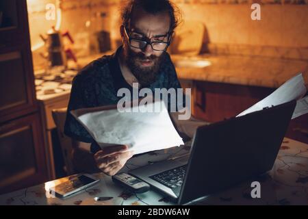 Stressato giovane hippster lunghi capelli bearded uomo sta controllando la fattura in kitchen.Invoice e finanza. Foto Stock