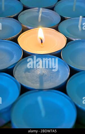 Primo piano delle luci del tè con una accesa. Una singola fiamma calda arancione tra candele in ombra blu. Natale, lutto o condoglienza concetto. Foto Stock