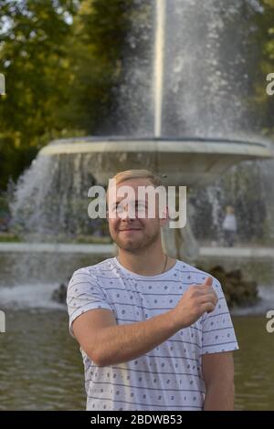 un giovane biondo in una maglietta leggera lancia una moneta nella fontana per una buona fortuna Foto Stock