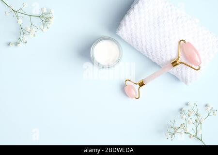 Rullo viso in quarzo rosa per trattamenti estetici con crema idratante, asciugamano e fiori su sfondo blu. Disposizione piatta, vista dall'alto, copia Foto Stock