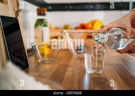 Le mani della donna versano l'acqua in un bicchiere nella sua cucina di casa Foto Stock