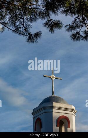 La croce bianca della chiesa ortodossa del villaggio di Fanari, regione di Xanthi, Grecia settentrionale, campanile vista parziale contro cielo blu con sovrastanti, incorniciato da rami di pino Foto Stock