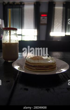 Pancake con uova e caffè in un vaso con paglia gialla su tavolo di legno nero e con sfondo non focalizzato. Foto Stock