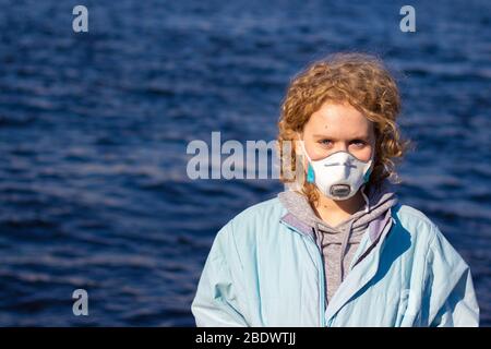 Una ragazza in una maschera medica protettiva sul suo viso guarda la fotocamera con uno sfondo di acqua blu. Femmina con capelli biondi ricci. Spazio di copia Foto Stock