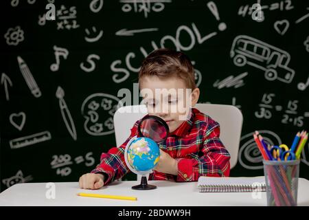 Carino ragazzo intelligente è seduto a una scrivania con lente di ingrandimento in mano. Bambino sta leggendo un libro con una lavagna su uno sfondo. Pronto per la scuola. Indietro Foto Stock