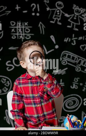 Carino ragazzo intelligente è seduto a una scrivania con lente di ingrandimento in mano. Bambino sta leggendo un libro con una lavagna su uno sfondo. Pronto per la scuola. Indietro Foto Stock