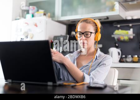 Freelance femmina nel suo casual home abbigliamento lavorando in remoto dal suo tavolo da pranzo al mattino. Cucina di casa in background. Foto Stock