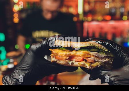 Le mani della donna con guanti di gomma nera stanno tenendo l'hamburger nero succoso con tagliatelle di carne, lattuga, pomodoro, formaggio grattugiato e cetriolo marinato. cor Foto Stock